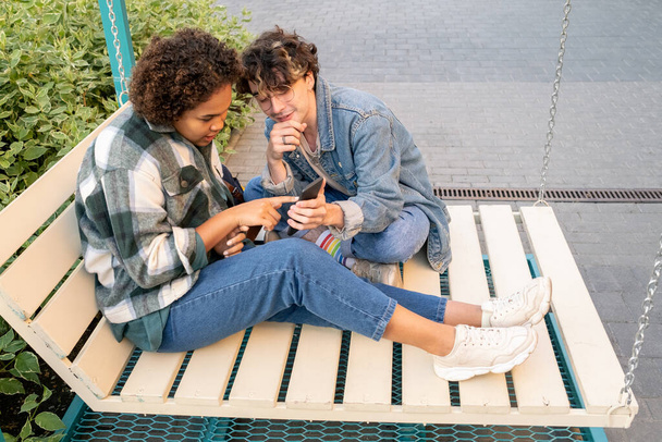 Two teenagers watching video in smartphone while sitting on swings - 写真・画像