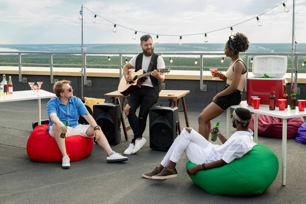 Group of restful friends with drinks listening to bearded man playing guitar - Fotó, kép