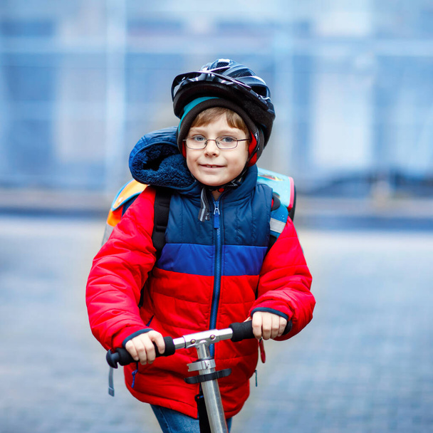 lindo niño de la escuela montado en scooter camino a la escuela primaria. Niño con casco de seguridad, bolso escolar en otoño lluvioso mañana fría. Tráfico en la ciudad y escolares. - Foto, imagen
