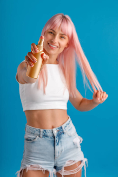 Jovem alegre com cabelo rosa sorrindo e mostrando spray hidratante para a câmera enquanto estava isolado sobre o fundo do estúdio azul - Foto, Imagem