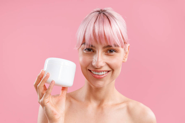 Retrato de mujer sonriente con cabello rosa sosteniendo tarro blanco con crema corporal nutritiva después de la ducha aislada sobre fondo rosa - Foto, Imagen