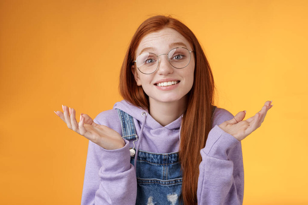 Well no big deal. Attractive silly redhead hipster girl wearing glasses smiling awkward unaware spread hands sideways shrugging confused grinning questioned uncertain what tell, orange background - Фото, изображение