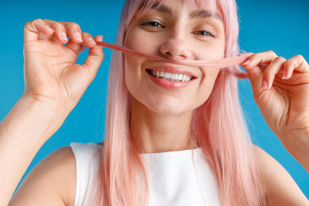 Close up portrait of happy young woman with natural long pink dyed hair holding a strand of hair as a moustache and smiling at camera, posing isolated over blue studio background - Фото, зображення