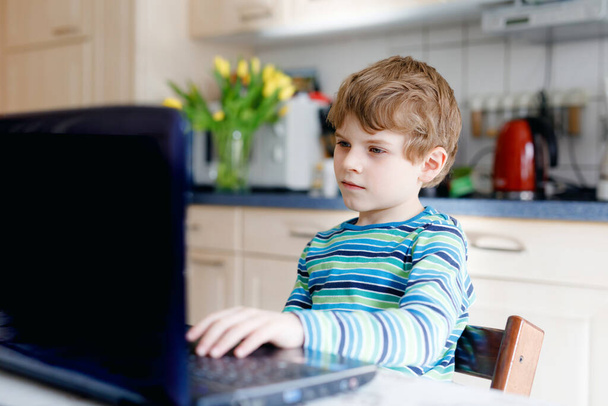 Παιδί αγόρι μαθαίνει στο σπίτι σε φορητό υπολογιστή για το σχολείο. Παιδί του δημοτικού σχολείου κάνει την εργασία και χρησιμοποιώντας σημειωματάριο και σύγχρονες συσκευές. Σπιτική σχολική ιδέα. Κάνοντας ένα μάθημα βίντεο με το δάσκαλο. - Φωτογραφία, εικόνα