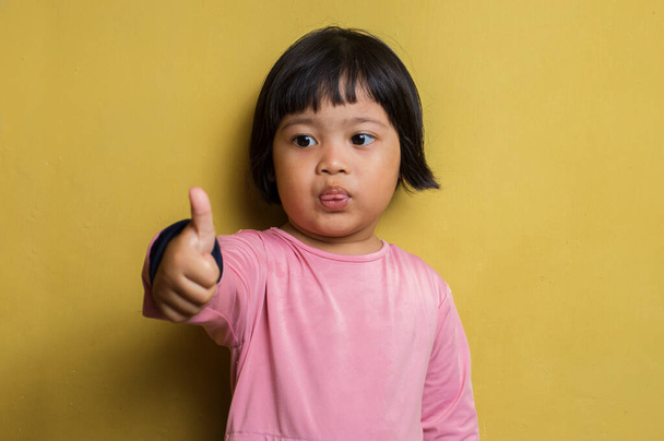 Asiatische kleine Mädchen mit ok Zeichen Geste tumb bis isoliert auf gelbem Hintergrund - Foto, Bild