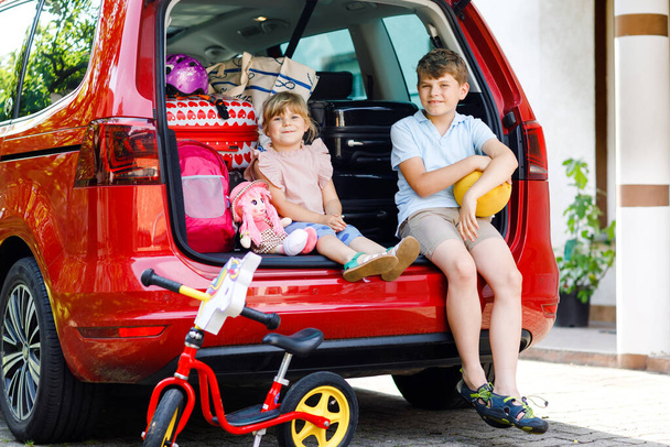 Due bambini, scolaro e bambina in età prescolare seduti nel bagagliaio dell'auto prima di partire per le vacanze estive con i genitori. Bambini felici, fratelli, fratelli e sorelle con valigie e giocattoli in viaggio - Foto, immagini