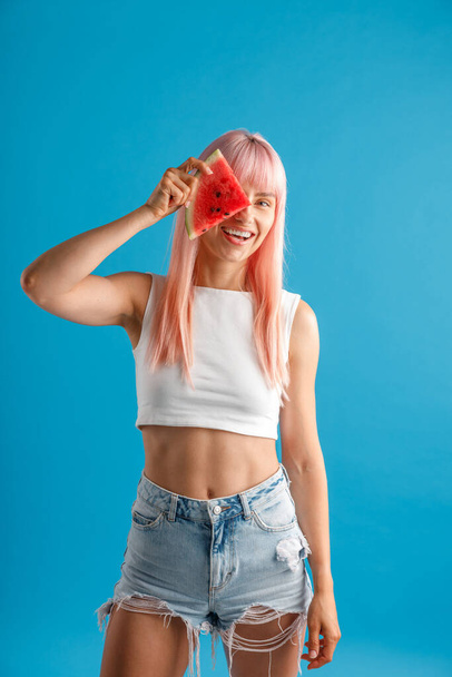 Lächelnde junge Frau mit rosafarbenen Haaren, die eine Scheibe Wassermelone über ihr Auge hält und isoliert vor blauem Studiohintergrund steht - Foto, Bild