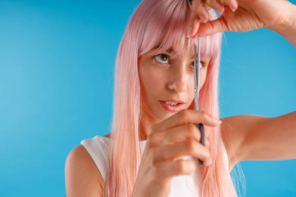 Retrato de una linda mujer joven con el pelo rosa cortando flecos a sí misma usando tijeras, de pie aislada sobre fondo de estudio azul - Foto, imagen