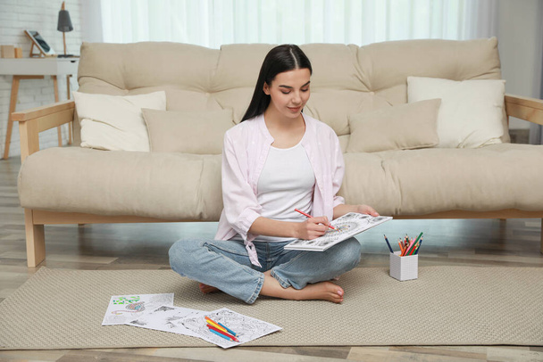 Young woman coloring antistress page near sofa in living room - Foto, Bild