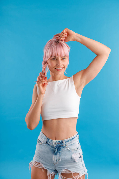 Jovencita alegre en ropa casual sonriendo a la cámara, jugando con su pelo rosa mientras posa aislada sobre fondo de estudio azul - Foto, imagen