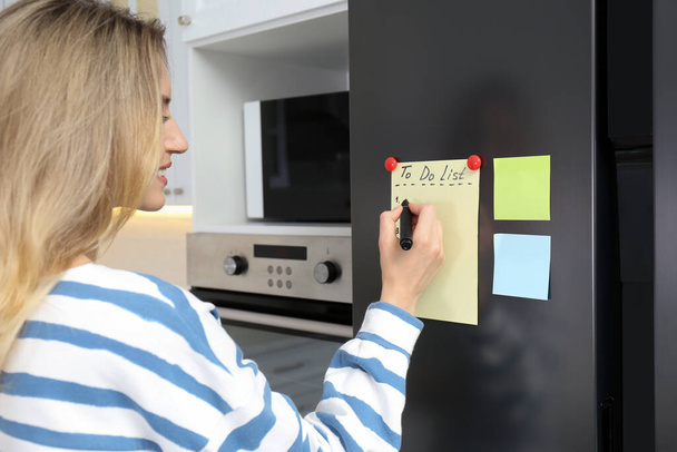 Young woman writing To do list on refrigerator door in kitchen - Zdjęcie, obraz