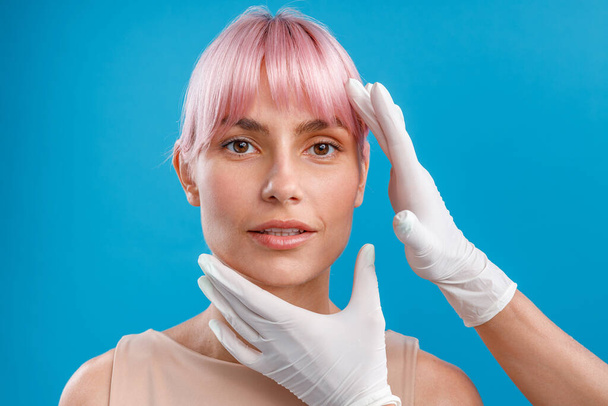 Hände der Kosmetikerin untersuchen das weibliche Gesicht, bevor sie Botox-Injektionen vor blauem Hintergrund verabreichen - Foto, Bild