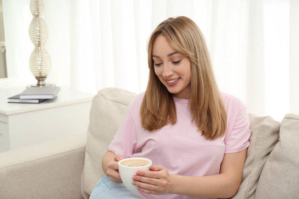 Beautiful young woman with cup of coffee relaxing on sofa at home - Foto, afbeelding