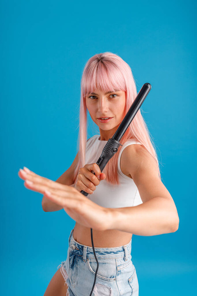 Playful young woman with pink hair looking at camera, making martial arts pose and holding curling iron, posing isolated over blue studio background - Fotoğraf, Görsel