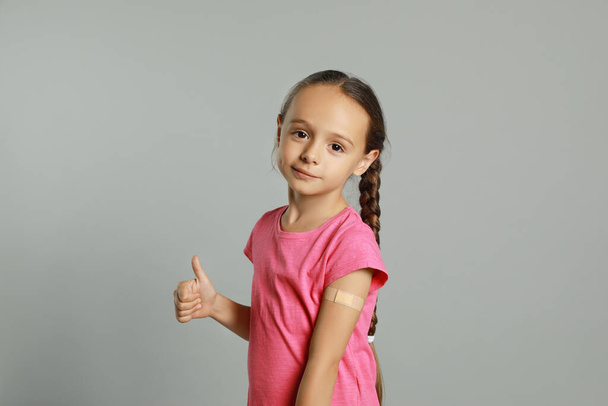 Vaccinated little girl with medical plaster on her arm showing thumb up against light grey background - Valokuva, kuva