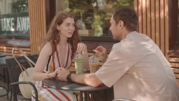 Середня тривалість молодої кавказької жінки, яка сидить за столом у кафе зі своїм чоловіком, пара тримається за руки, п'є з льодом, розмовляє і посміхається. - Кадри, відео
