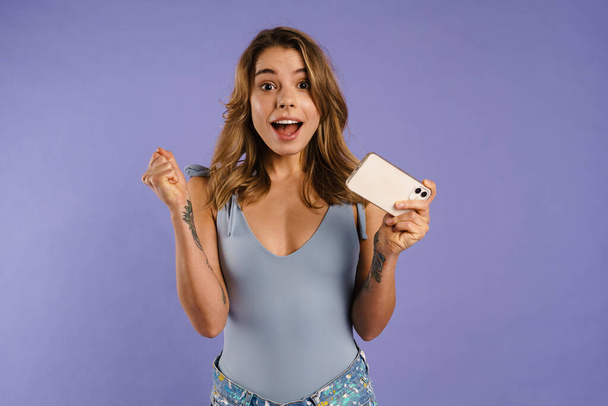 Young white woman rejoicing and holding cellphone isolated over purple background - Фото, изображение