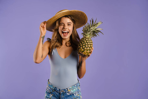 Young white woman in straw hat smiling and holding pineapple isolated over purple background - Фото, зображення