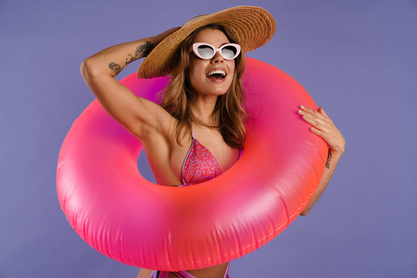 Young white woman in swimsuit and sunglasses holding rubber ring isolated over purple background - Foto, immagini