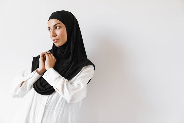 A concentrated muslim woman in hijab raising her arms and looking to the side while standing in the white studio - 写真・画像