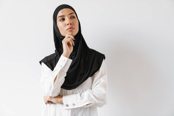 A pensive muslim woman with pierced nose in hijab thinking about something while standing in the white studio - Photo, image