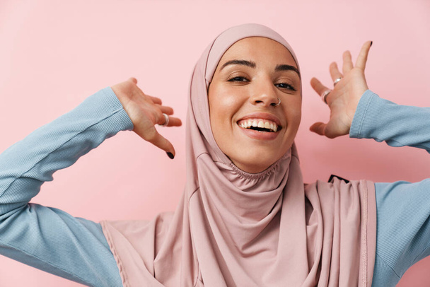 A portrait of the laughing muslim woman wearing pink hijab standing with raised arms in the pink studio - 写真・画像