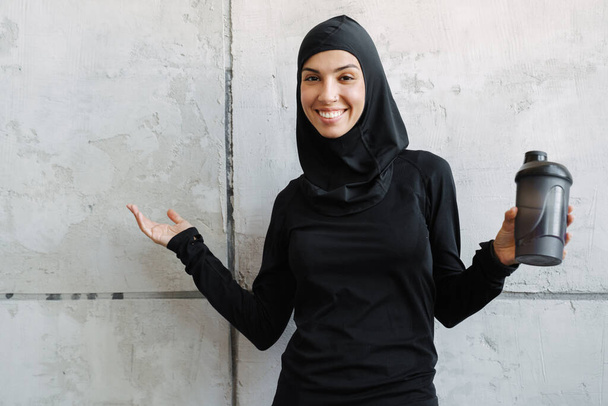 Young muslim woman in hijab smiling while holding water bottle and copyspace indoors - Fotoğraf, Görsel