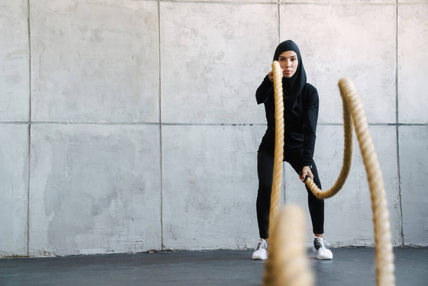 Young muslim woman in hijab working out with battle ropes indoors - Foto, Imagen