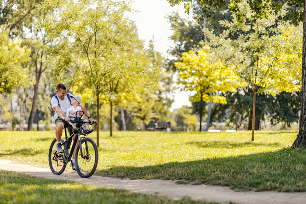 Otec a syn se sbližují v přírodě. Malý chlapec sedí v koši na kole, zatímco jeho otec projíždí lesem na kole. Krásný slunečný den v přírodě se poflakovat s rodinou - Fotografie, Obrázek