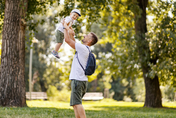 The family has fun in the park, escape to nature and a family active weekend. Father and son play in the woods dressed in the same casual clothes on a sunny day. Dad lifts the boy up and he giggles - Valokuva, kuva
