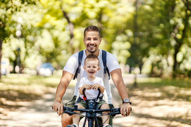 Vater und Sohn verbinden Zeit in der Natur. Ein kleiner Junge sitzt in einem Fahrradkorb, während sein Vater mit dem Fahrrad durch den Wald fährt. Schöner sonniger Tag in der Natur, um mit der Familie abzuhängen. Lächelnd - Foto, Bild