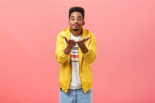 Blowing you sweet kisses. Cute and handsome stylish man with beard and afro hairstyle bending towards camera folding lips and smiling sending mwah with hands near breast over pink background - Photo, Image