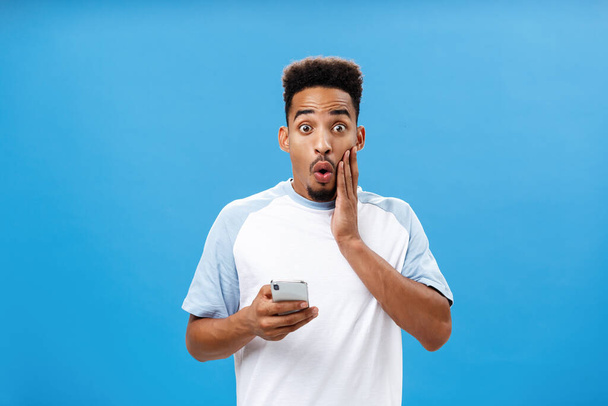 Waist-up shot of charmed intrigued handsome stylish dark-skinned male with afro haircut folding lips in wow or awesome expression holding cheek on face holding smartphone reacting to amazing news - Photo, Image