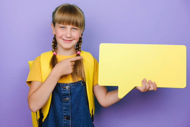 Portrait of cute positive caucasian school girl pointing on yellow speech bubble, wears backpack, posing isolated over purple color background in studio with copy space for promotion content. Mockup - Photo, Image
