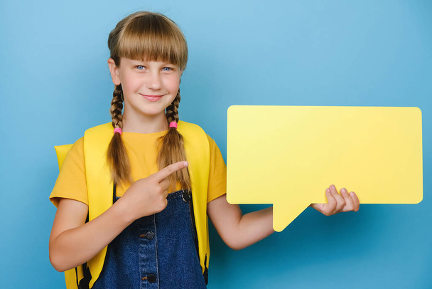 Portrait of cute happy little schoolgirl holding empty speech bubble and pointing finger, wears yellow backpack, posing isolated over blue studio background with copy space. Back to school concept - Fotografie, Obrázek
