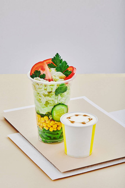 Takeaway food. Cappuccino coffee and salad with fresh vegetables in a plastic cup on a beige background. Space for text - Photo, Image