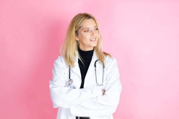 jonge blonde arts vrouw dragen stethoscoop staan over geïsoleerde roze achtergrond glimlachen en kijken naar de zijkant - Foto, afbeelding