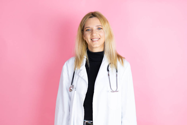 Νεαρή ξανθιά γιατρός γυναίκα φορώντας στηθοσκόπιο στέκεται πάνω από απομονωμένο ροζ φόντο χαμογελώντας και κοιτάζοντας την κάμερα - Φωτογραφία, εικόνα