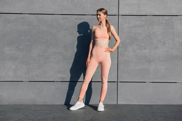 Young fitness woman with a beautiful body, in sports pink clothes, exercising outdoors, against the background of an urban gray wall, Female model in sportswear, exercising outdoors, Sports, Motivation - Fotoğraf, Görsel
