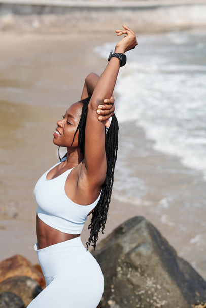 Lächelnde junge Frau genießt gute Rückenstreckung nach dem Training am Strand - Foto, Bild