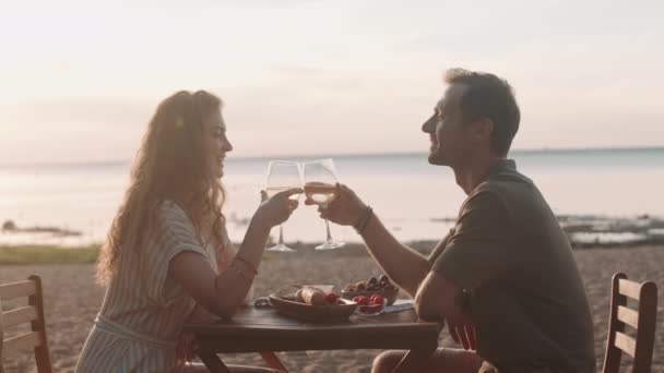 喜びのサイドビュー白人女性と男が砂の海岸のテーブルに座って、白ワインとロマンチックなディナーを持ち、楽しい会話をして - 映像、動画
