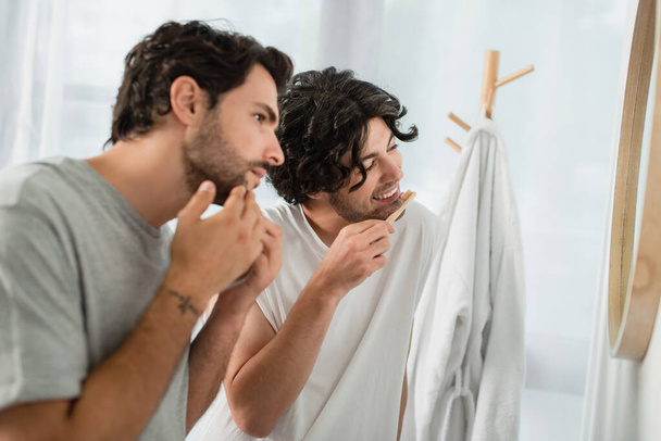 borrosa y tatuado gay hombre tocando cara mientras novio cepillarse los dientes y sonriendo en cuarto de baño  - Foto, Imagen