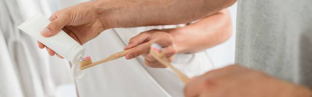 vista parcial del hombre apretando la pasta de dientes en el cepillo de dientes cerca de novio en el baño, pancarta - Foto, imagen