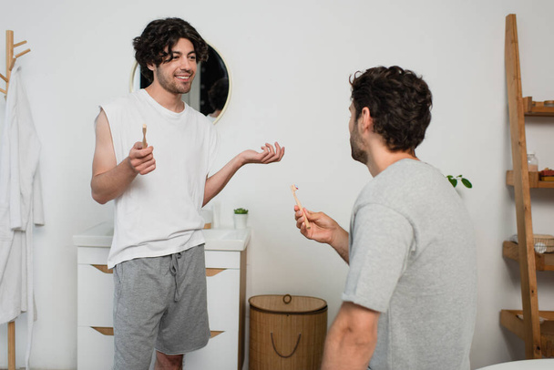 smiling gay couple holding toothbrushes while talking in bathroom - 写真・画像