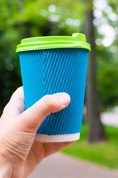 Γυαλί με καφέ σε γυναικείο χέρι στο φόντο ενός πάρκου, πράσινο γρασίδι και δέντρα. - Φωτογραφία, εικόνα