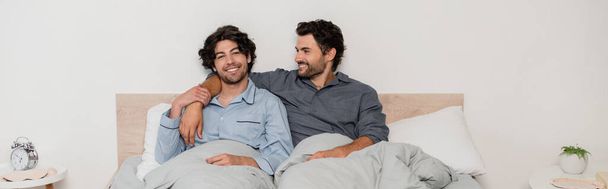 fröhliches homosexuelles Paar lächelt, während es im Bett chillt, Banner - Foto, Bild