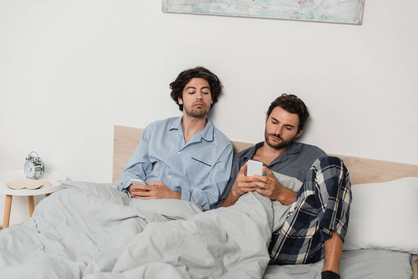 άντρας που κοιτάει το κινητό ενώ ο φίλος της μιλάει στο κρεβάτι - Φωτογραφία, εικόνα
