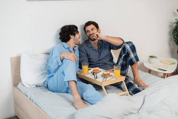 cheerful gay couple looking at each other near tray with breakfast  - Foto, afbeelding