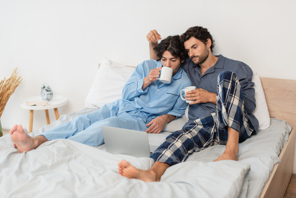 gay paar holding cups van thee en kijken film op laptop in bed - Foto, afbeelding