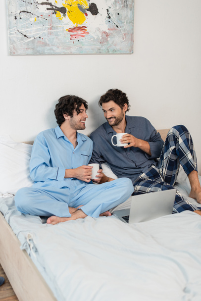 happy gay couple holding cups of tea near laptop in bed - Фото, зображення
